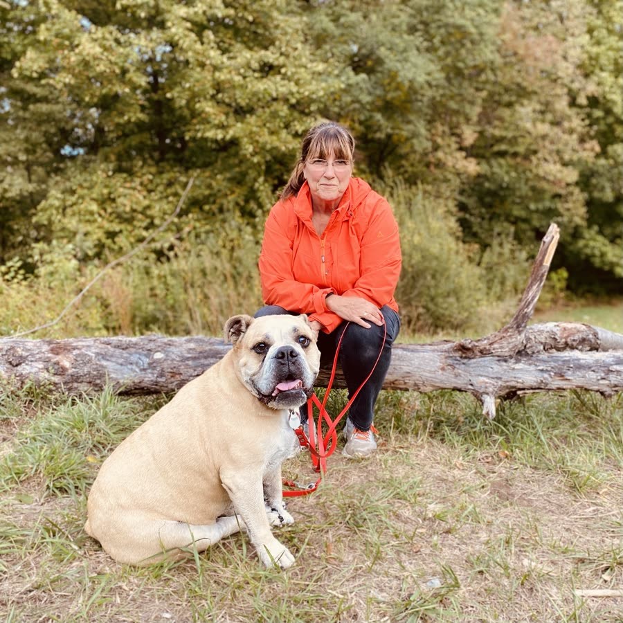 Erika Siefert sitzt auf einem Baumstamm im Grünen, ihre Bulldogge sitzt an der roten Leine vor ihr